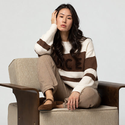 Femme assise dans un fauteuil portant le pull en cachemire PROUST rayé écru et chocolat, en précommande MAX&MOI en édition limitée. Le pull est porté avec le pantalon BLIZZARD en laine et cachemire. Collection Automne-Hiver 26  MAX&MOI
